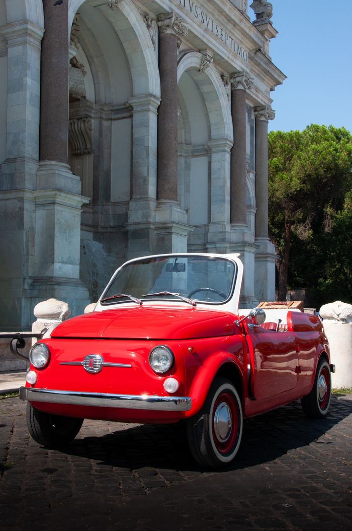 showroom Classic red Fiat 500 cabriolet parked by an ancient Italian archway.
